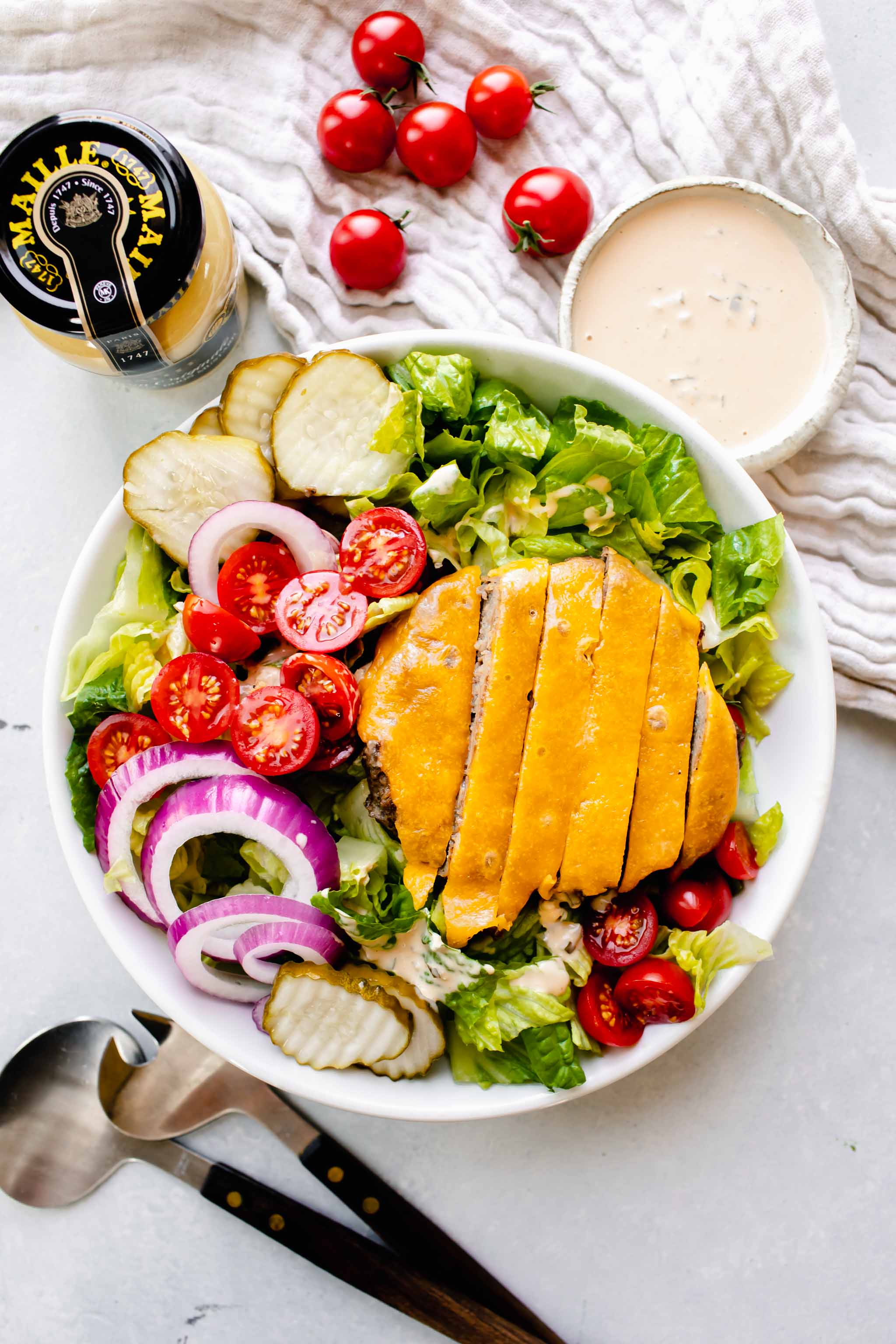 Overhead shot of bowl of cheeseburger salad next to jar of mustard and bowl of special sauce.