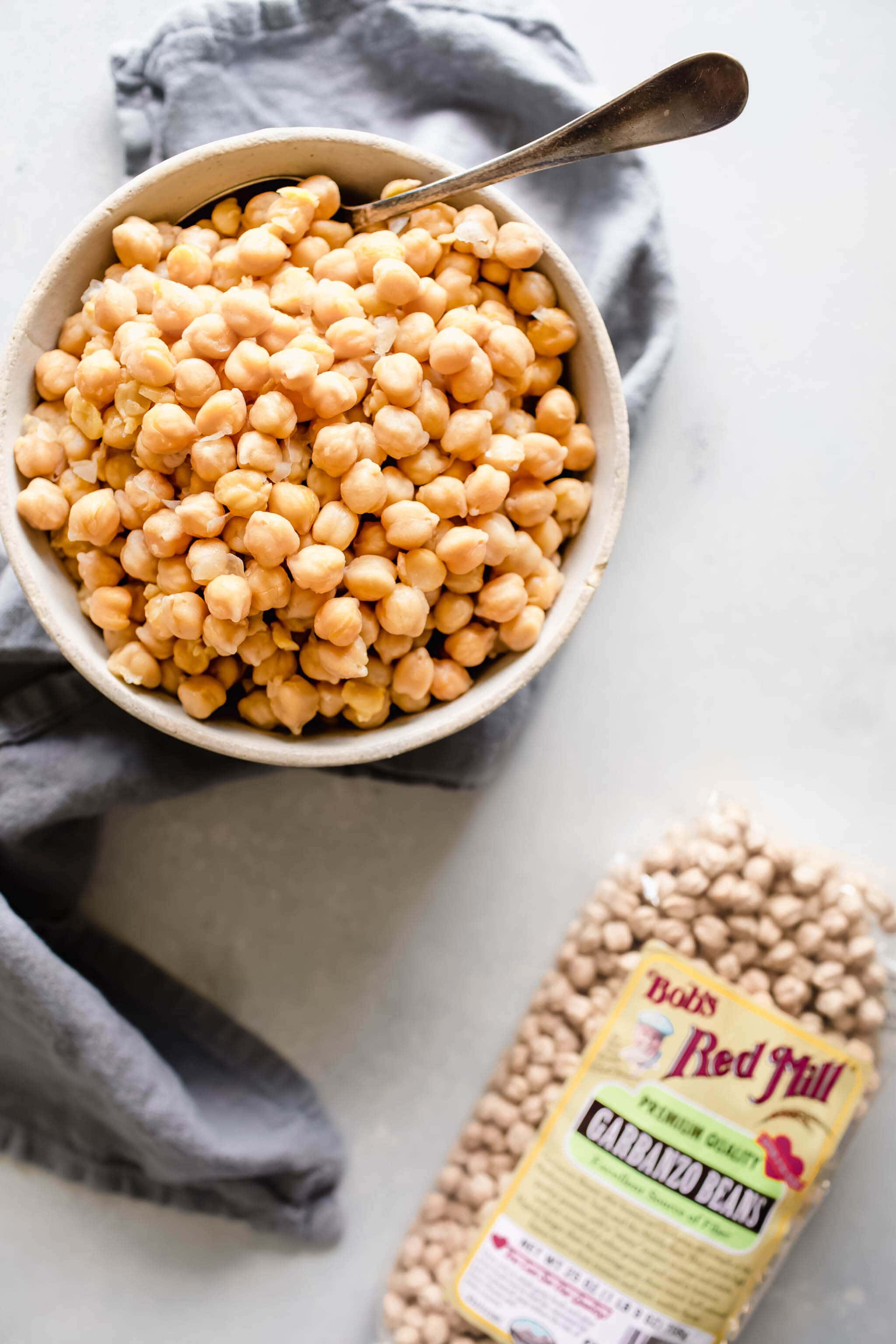 Bowl of cooked chickpeas with spoon next to bag of uncooked chickpeas.