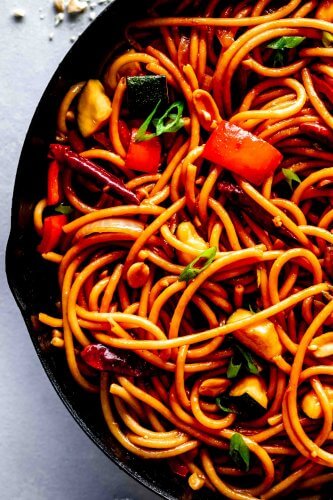 Kung Pao noodles in cast iron skillet on grey counter flecked with chopped peanuts.