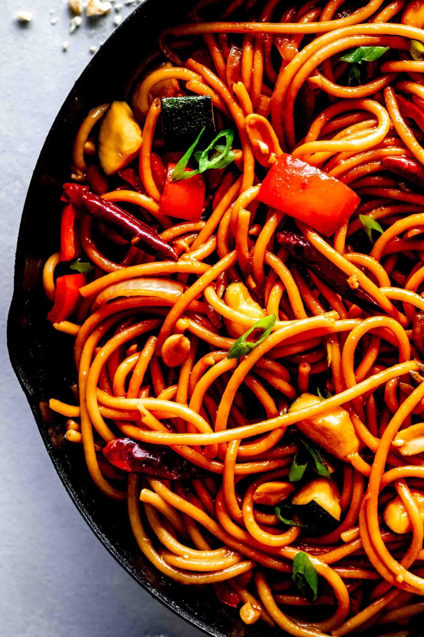 Kung Pao noodles in cast iron skillet on grey counter flecked with chopped peanuts.