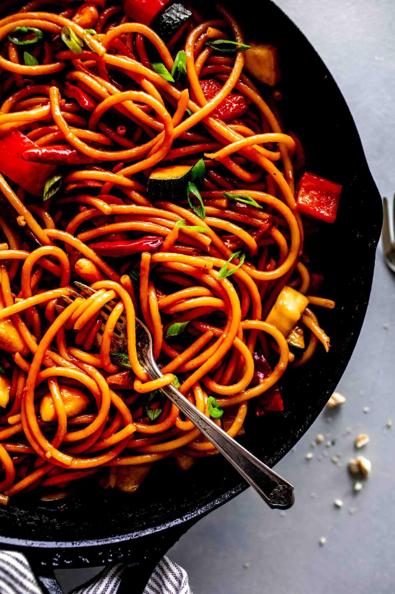 Kung Pao noodles in cast iron skillet on grey counter flecked with chopped peanuts and fork in skillet with pasta twirled around it. 