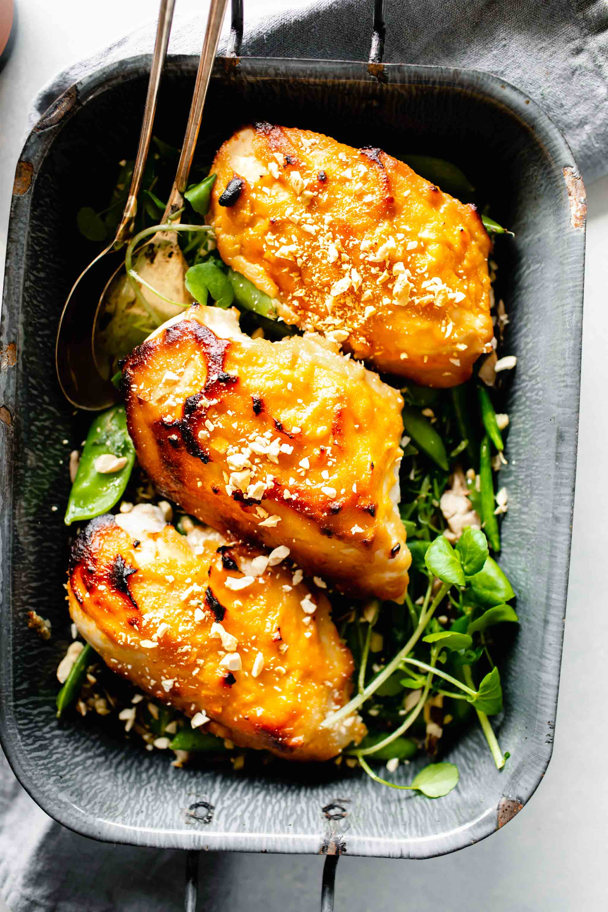 Overhead shot of three chicken breasts glazed with miso in grey pan served over pea pods.