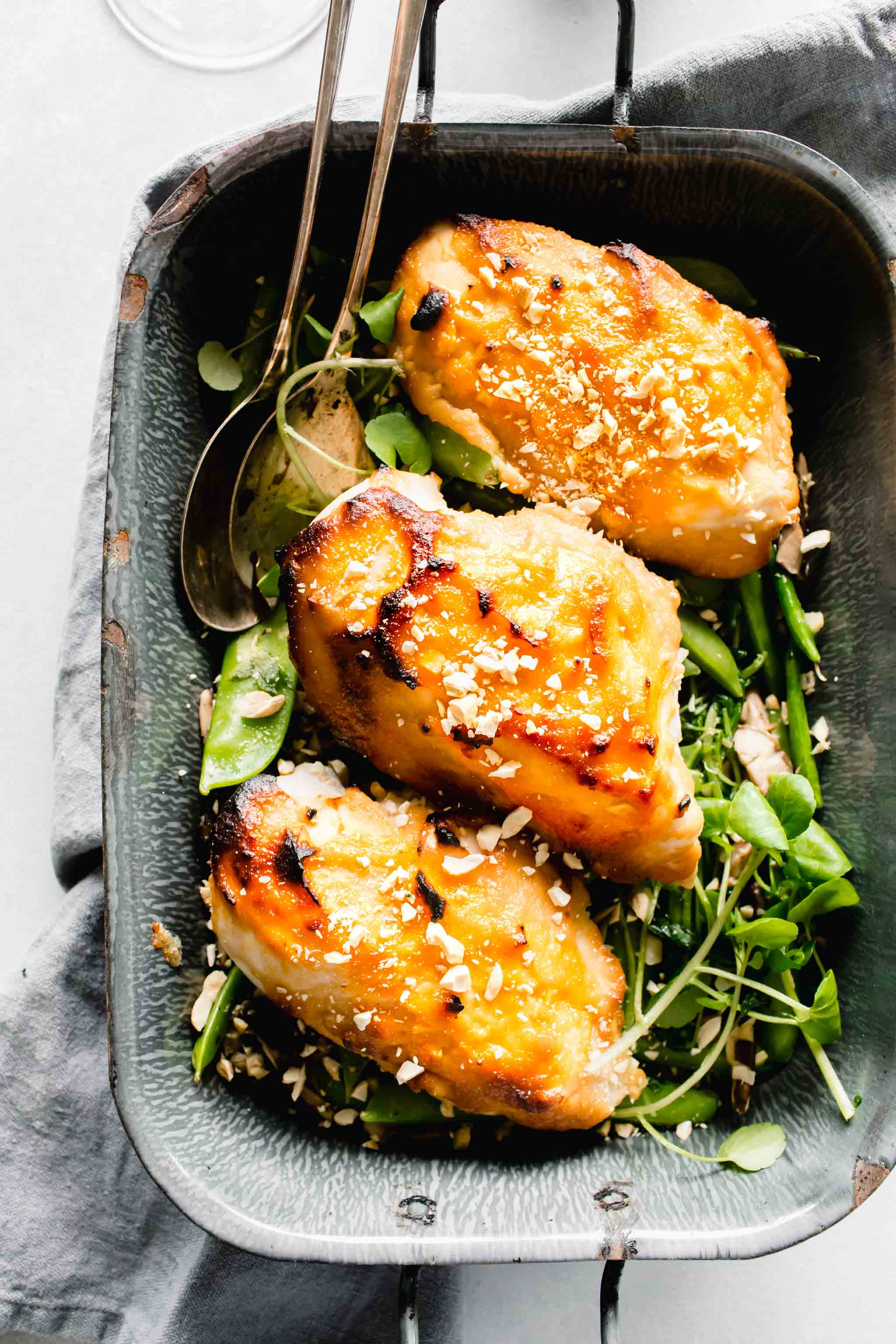 Overhead shot of three chicken breasts glazed with miso in grey pan served over pea pods.