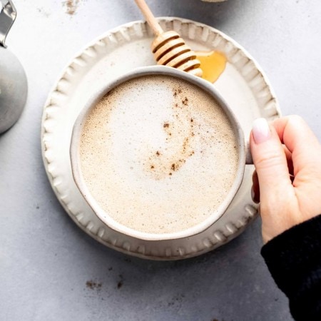 Hand holding cream colored mug of honey cardamom latte next to small dish of honey and honey wand.