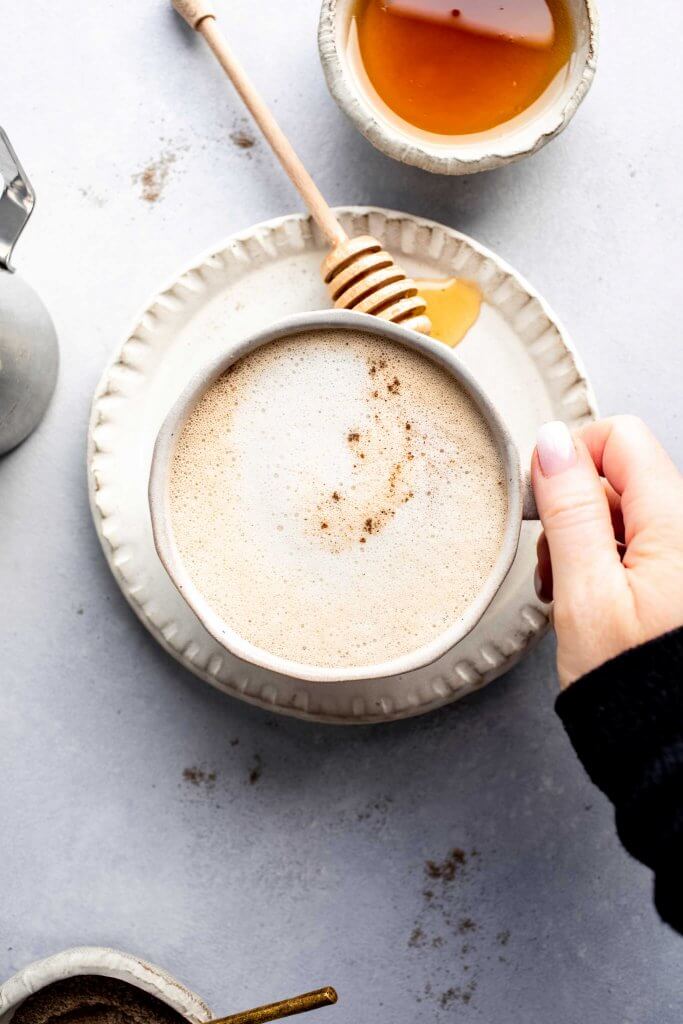 Hand holding cream colored mug of honey cardamom latte next to small dish of honey and honey wand.