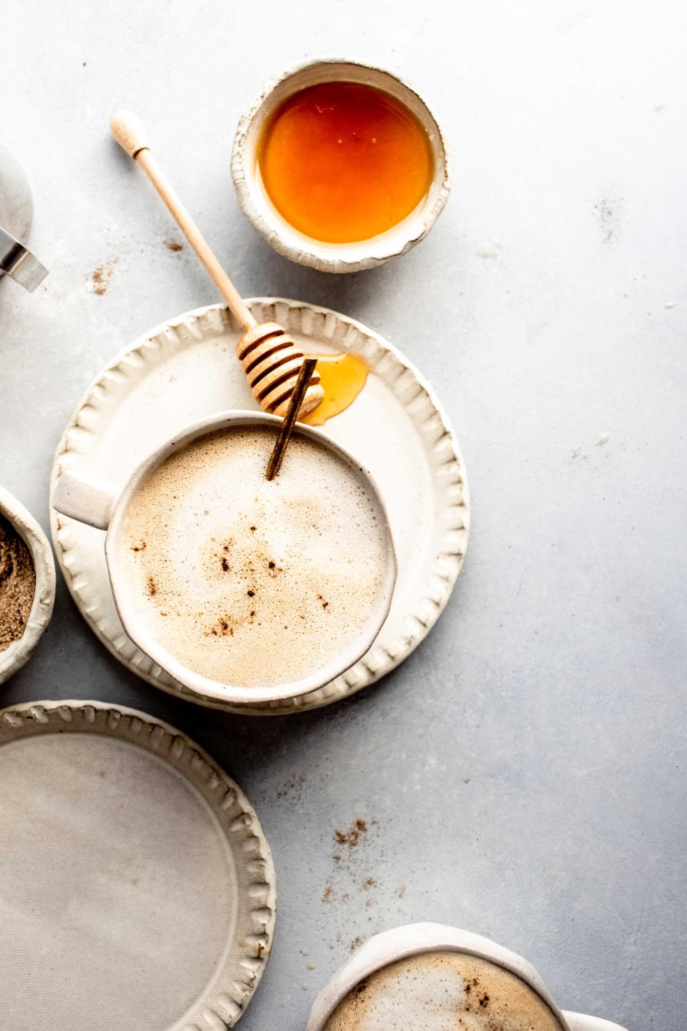 Two mugs of cardamom latte next to pinch bowl of cardamom and small bowl of honey.