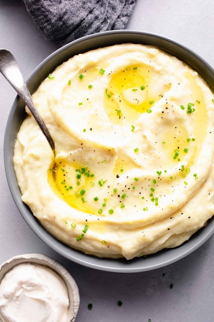 Overhead close up of instant pot mashed potatoes in grey bowl with two pats of melted butter & chives next to bowl of sour cream.