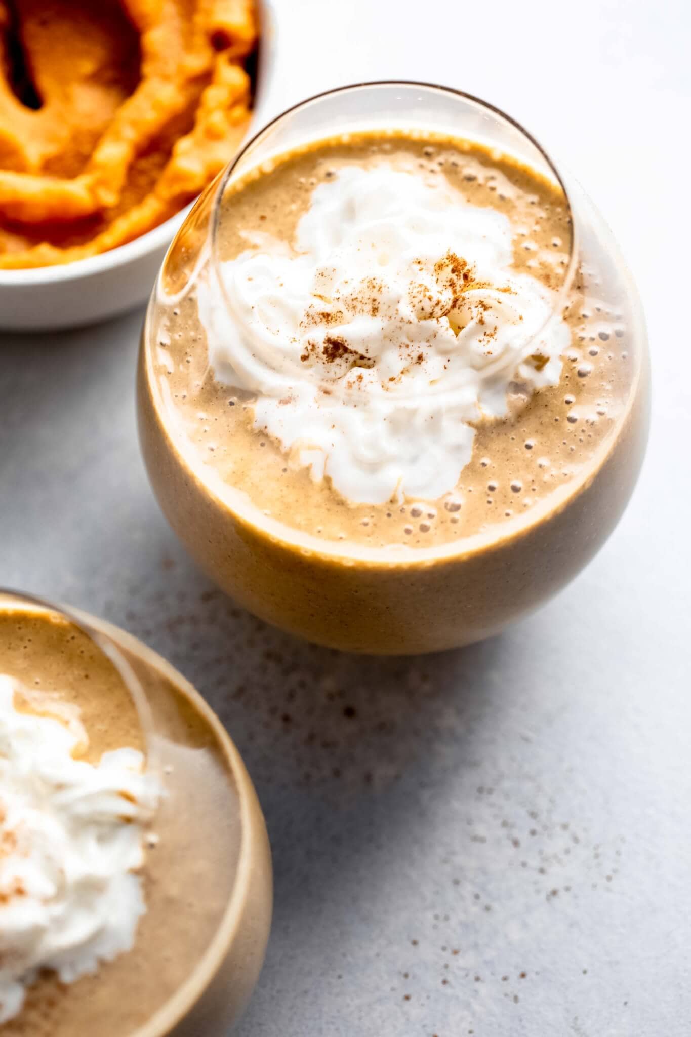 Two pumpkin smoothies topped with whipped cream next to bowl of pumpkin. 