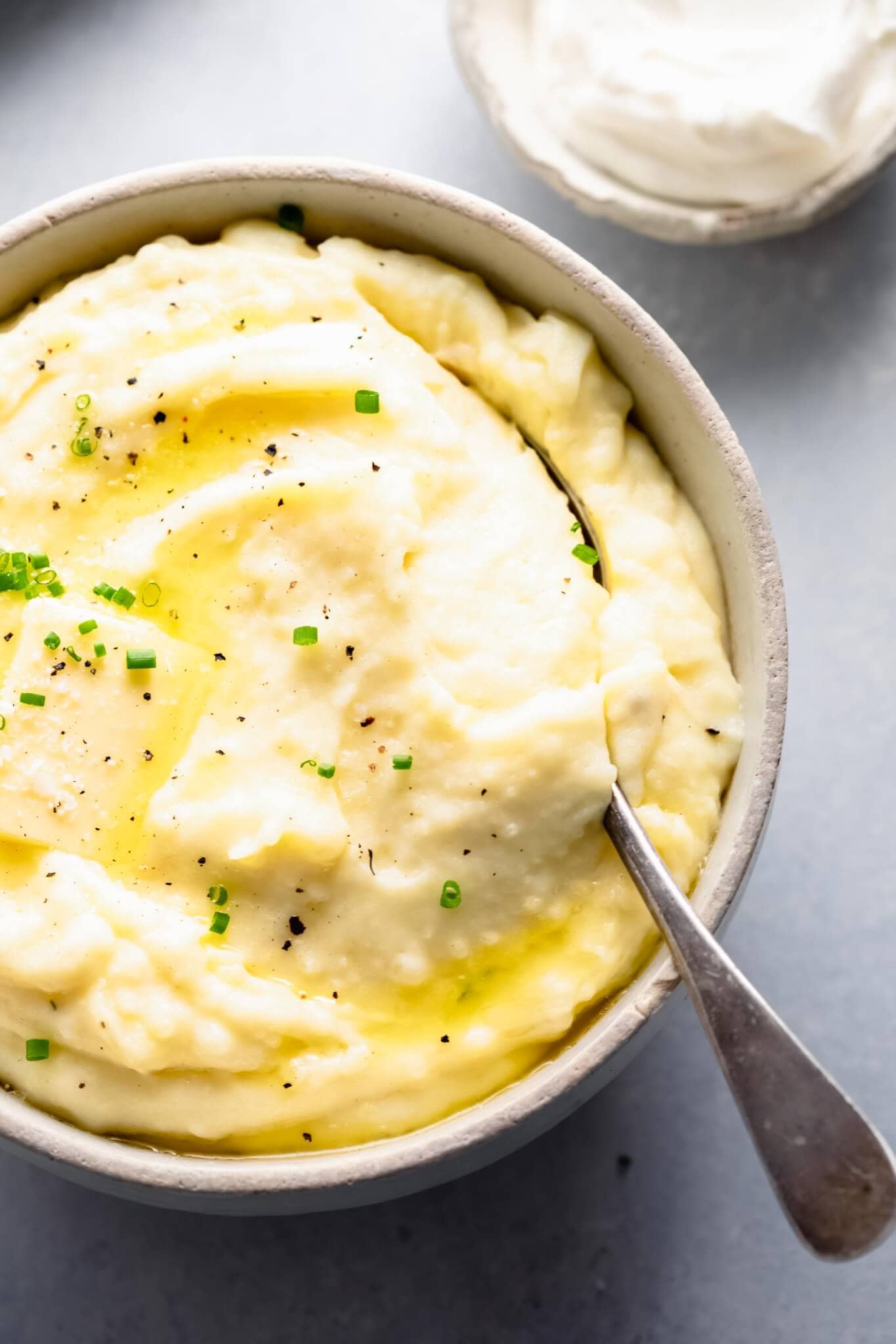 Overhead shot of bowl of mashed potatoes topped with a pat of butter, next to bowl of sour cream.