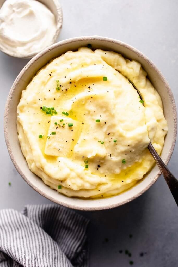 Overhead shot of bowl of mashed potatoes topped with a pat of butter, next to bowl of sour cream.