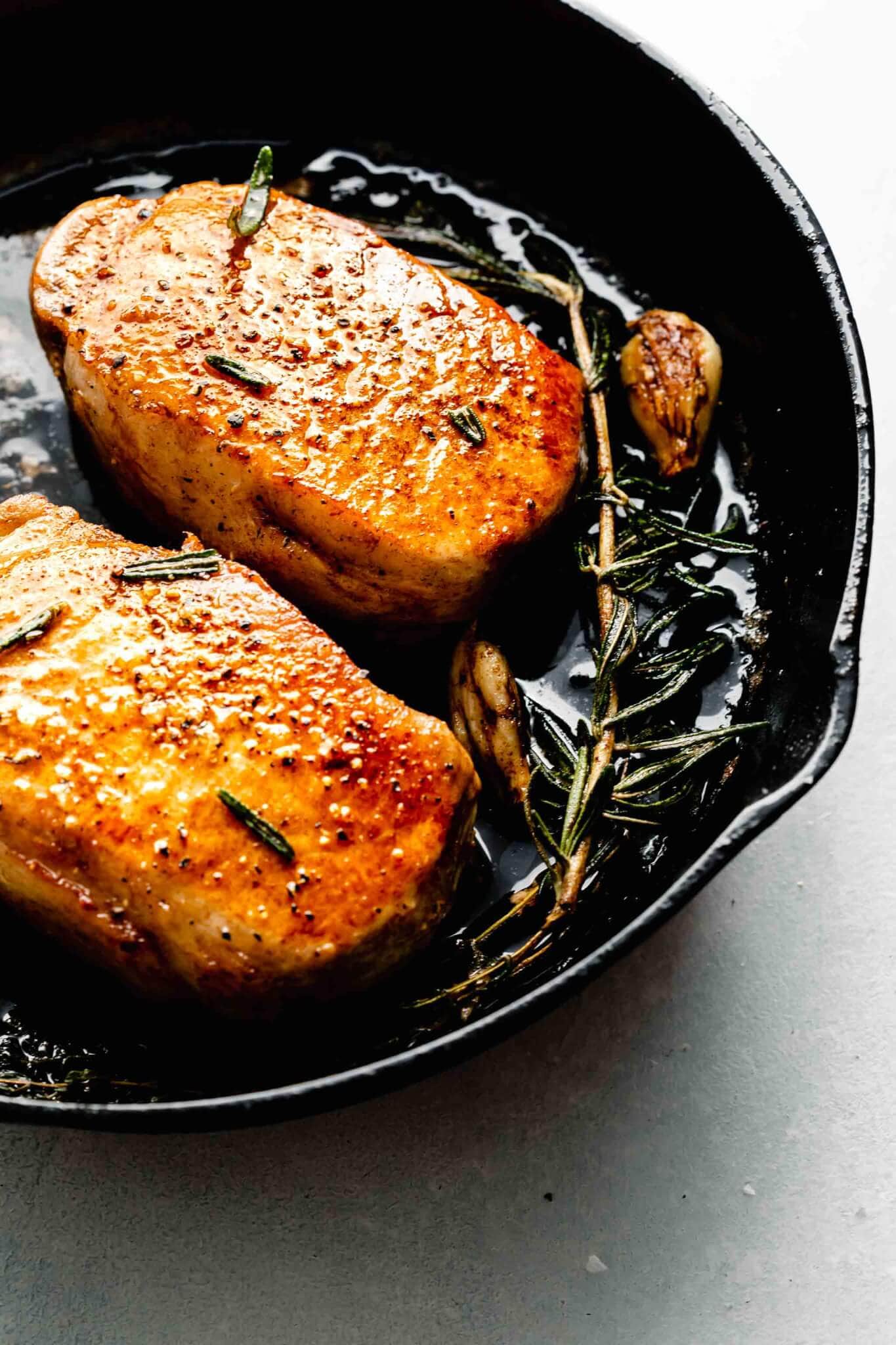 Two cooked sous vide pork chops in cast iron skillet with sprig of rosemary and garlic clove.