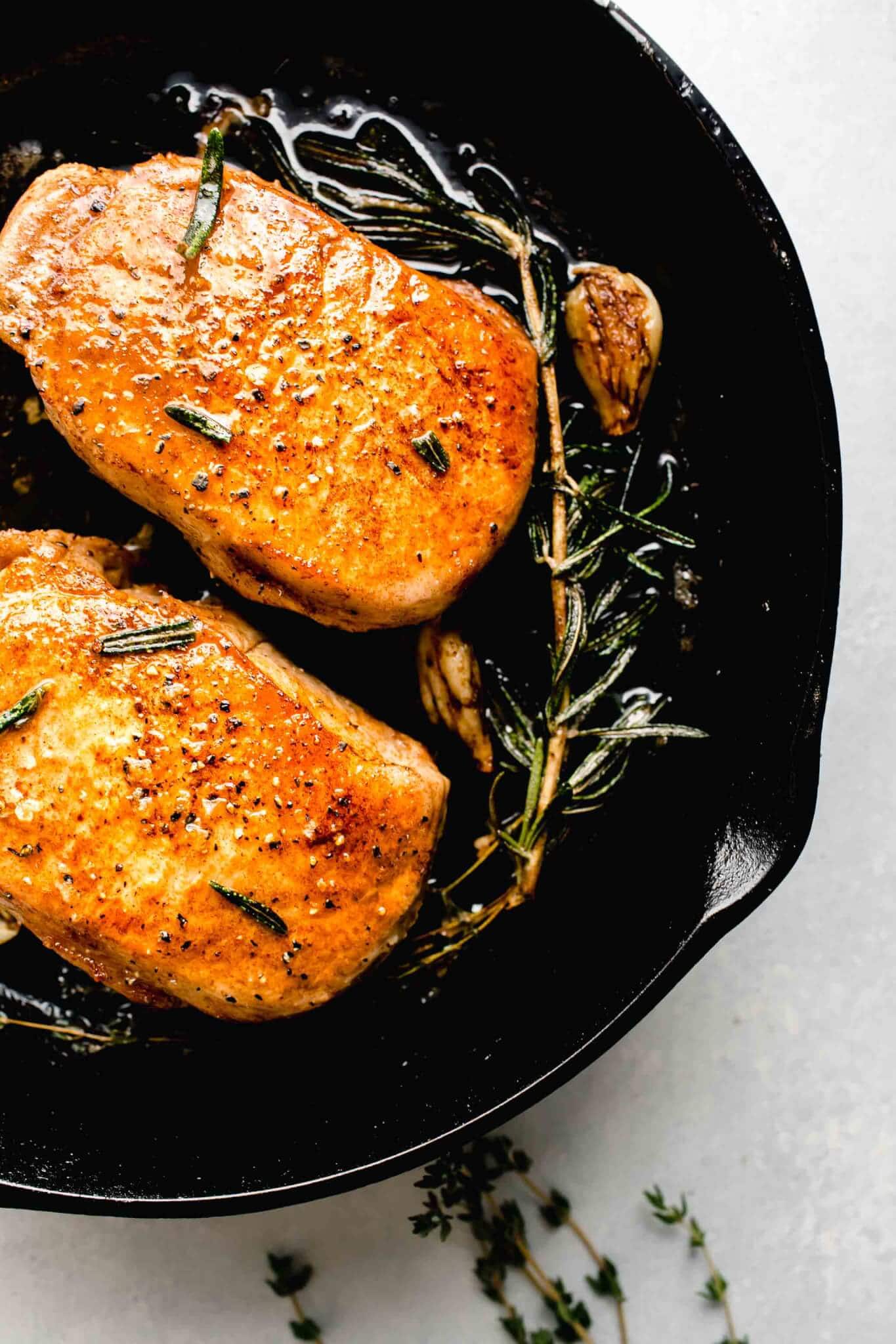 Two cooked sous vide pork chops in cast iron skillet with sprig of rosemary and garlic clove.