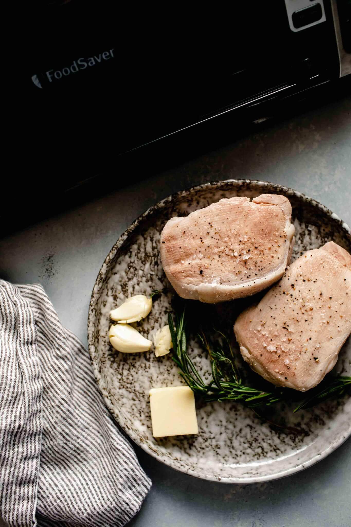 Two pork chops on a grey plate next to pat of butter and garlic cloves. Finished cooking sous vide ready to be seared.