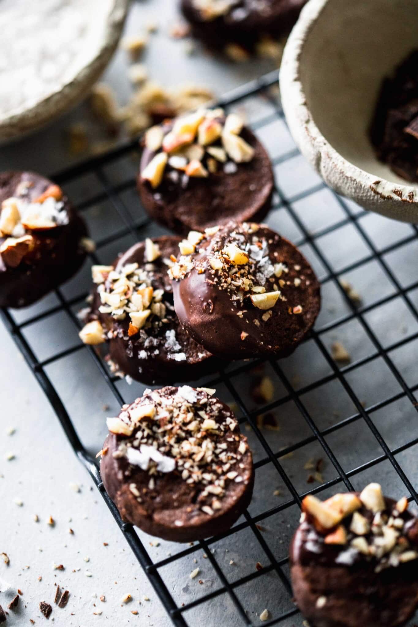 Chocolate shortbread cookies on cooling rack next to bowls of chocolate and sea salt.