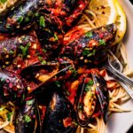 Close up of mussels in marinara sauce served over spaghetti with lemon wedges.