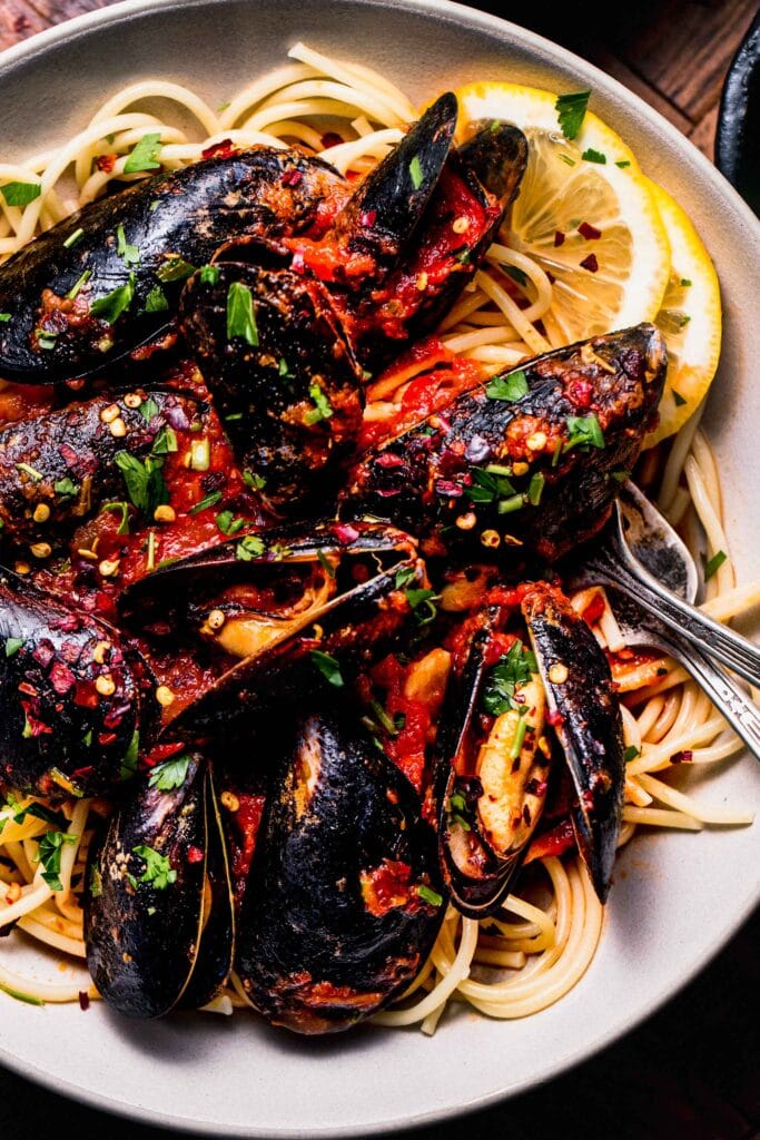 Close up of mussels in marinara sauce served over spaghetti with lemon wedges.