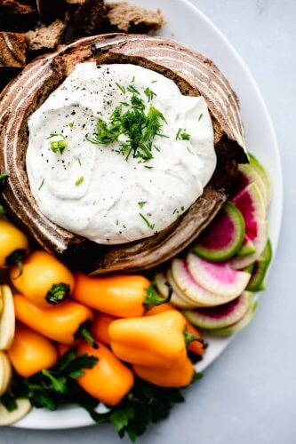 Overhead shot of greek yogurt ranch dip in bread bowl next to small orange peppers and watermelon radish.