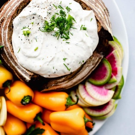 Overhead shot of greek yogurt ranch dip in bread bowl next to small orange peppers and watermelon radish.