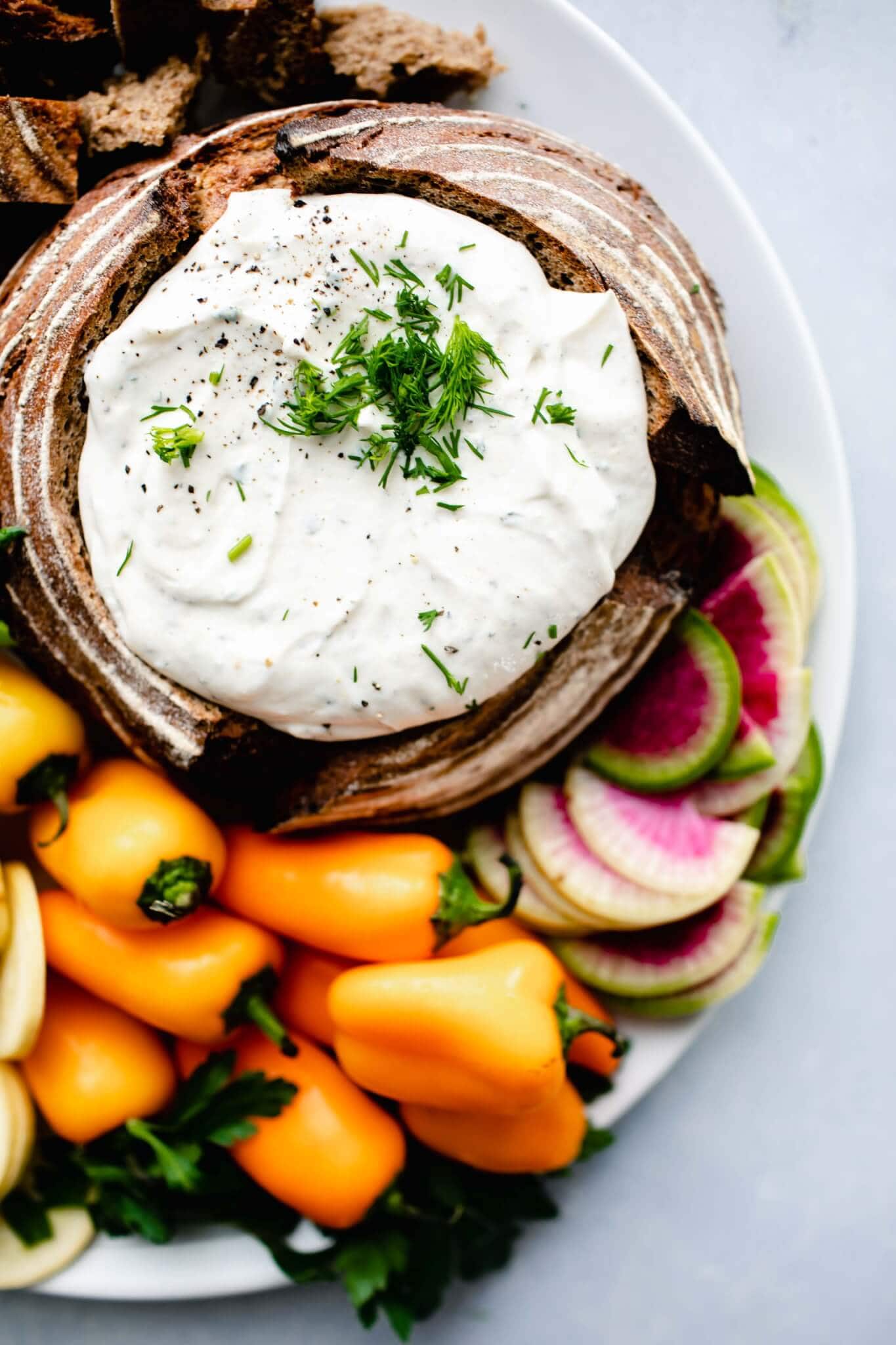 Overhead shot of greek yogurt ranch dip in bread bowl next to small orange peppers and watermelon radish.