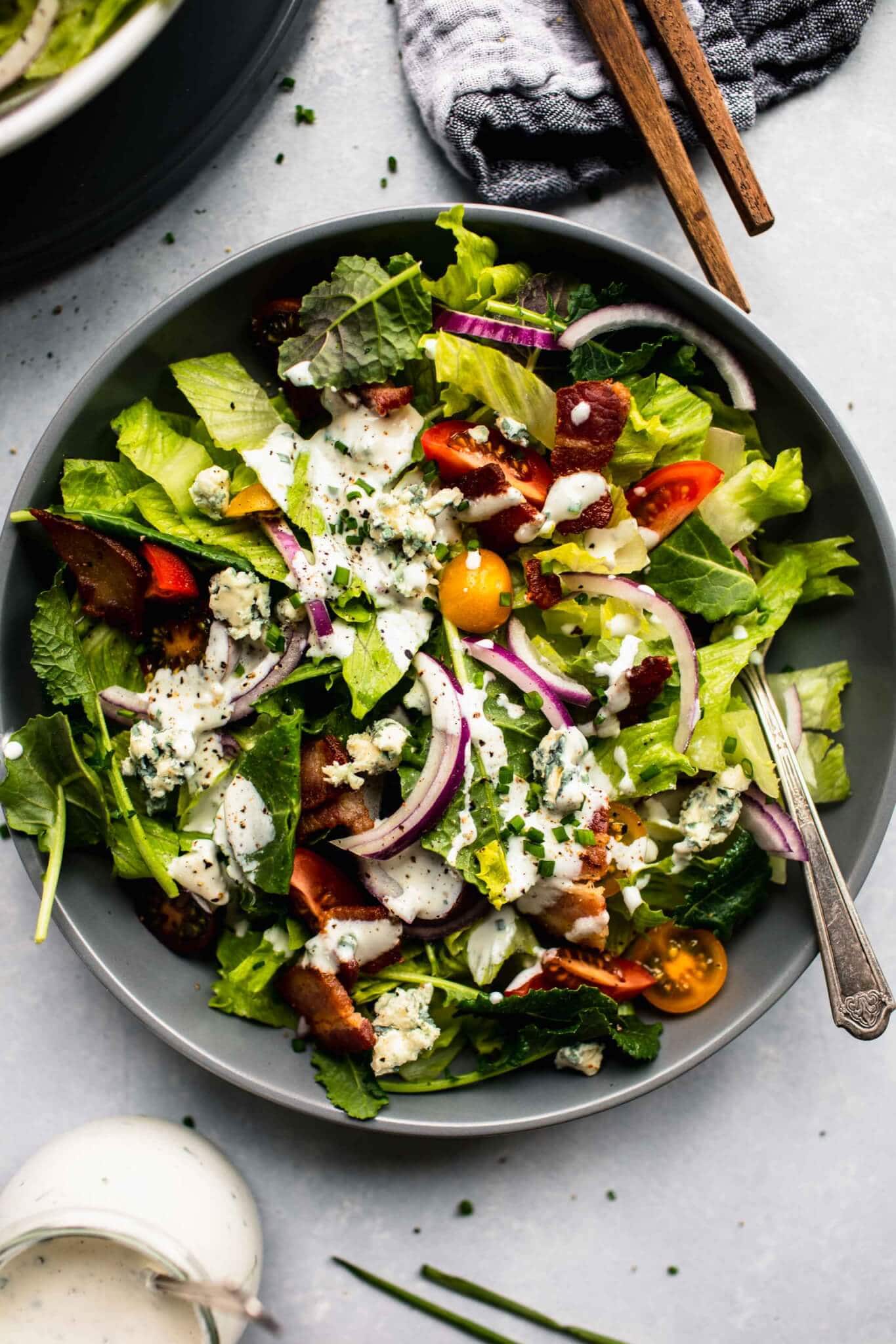 Green salad in grey bowl topped with red onions, tomatoes and blue cheese dressing, with serving utensils and small jar of dressing.