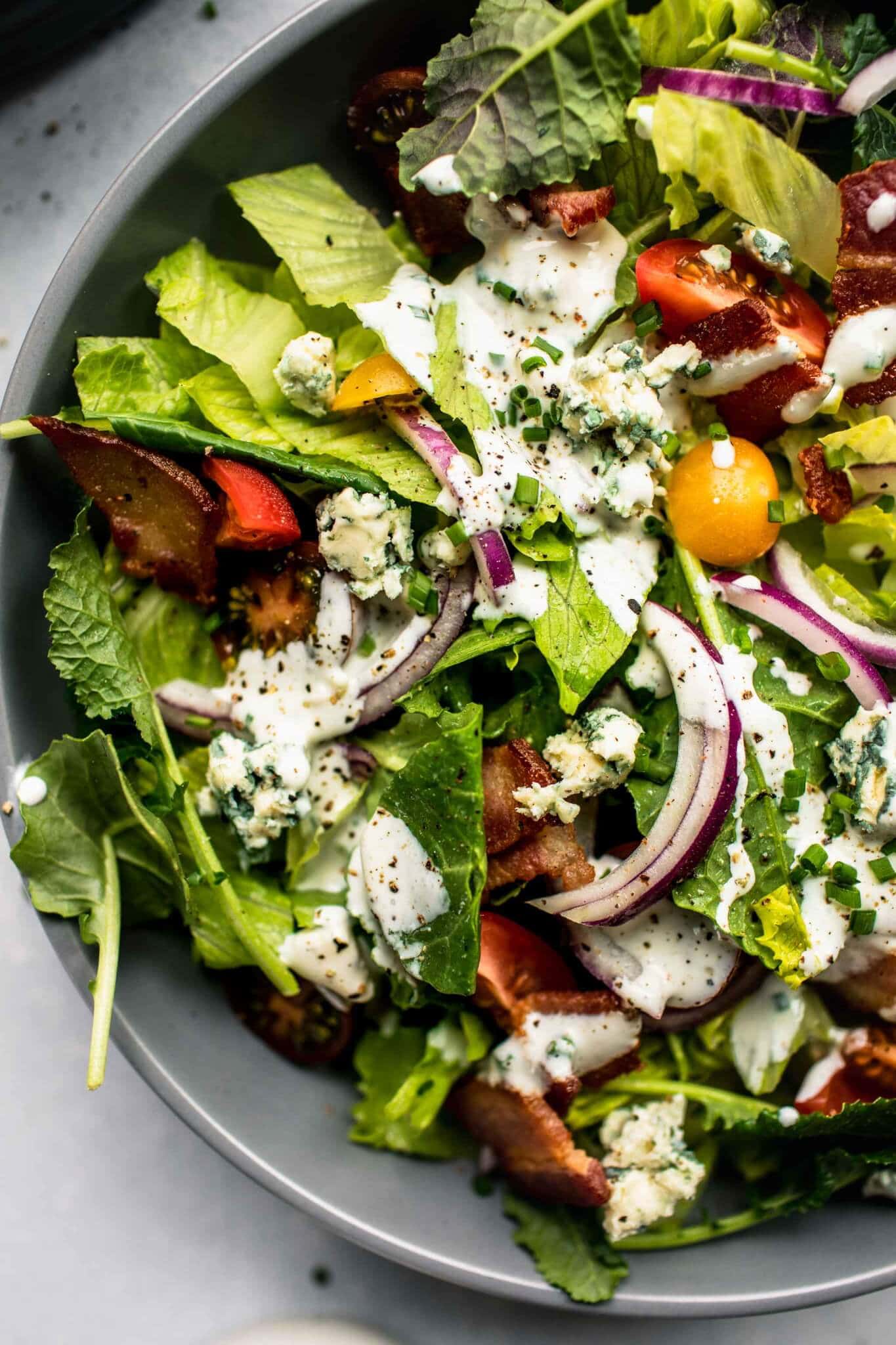 Green salad in grey bowl topped with red onions, tomatoes and blue cheese dressing.