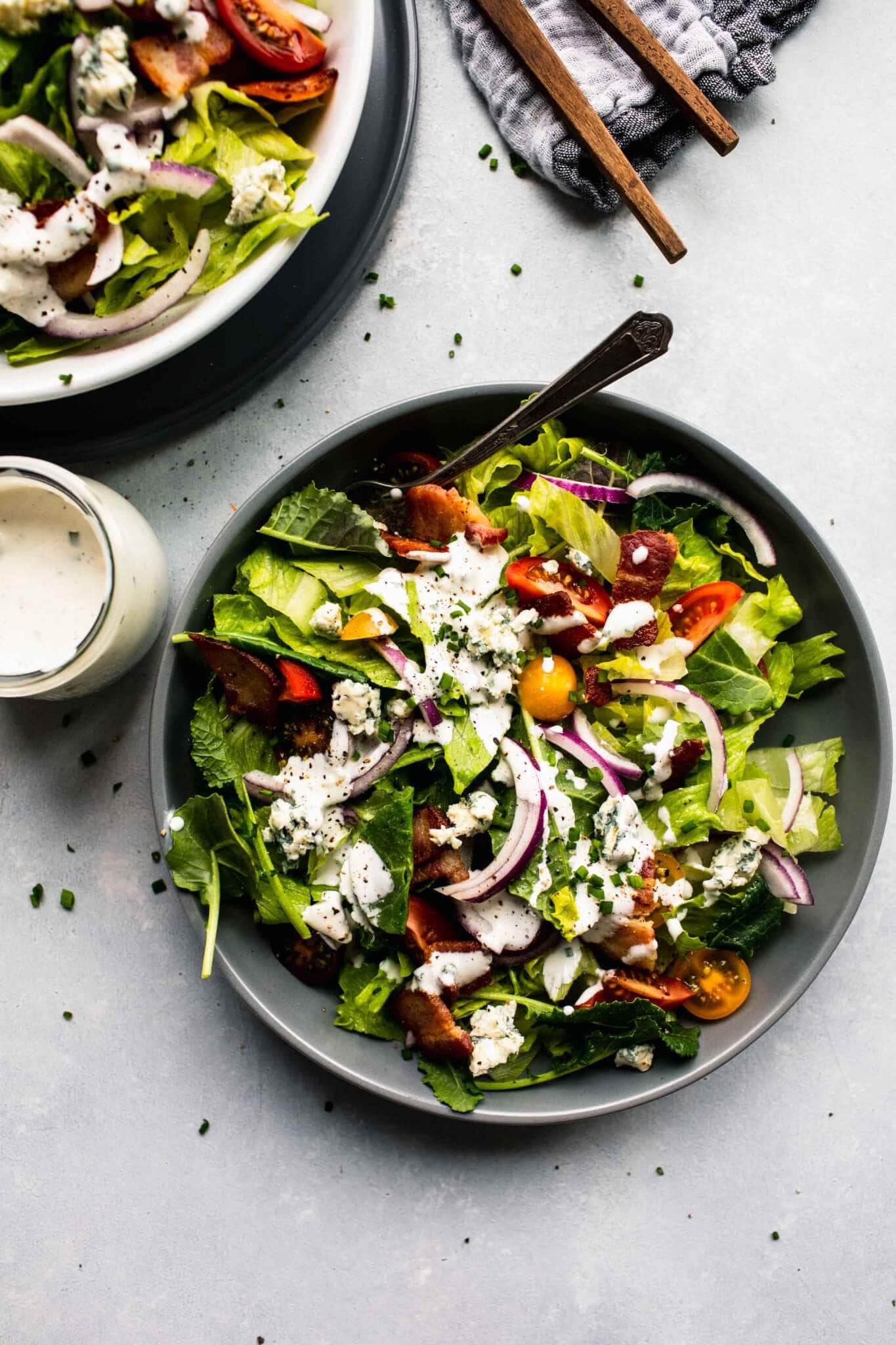 Green salad in grey bowl topped with red onions, tomatoes and blue cheese dressing, with serving utensils and small jar of dressing.