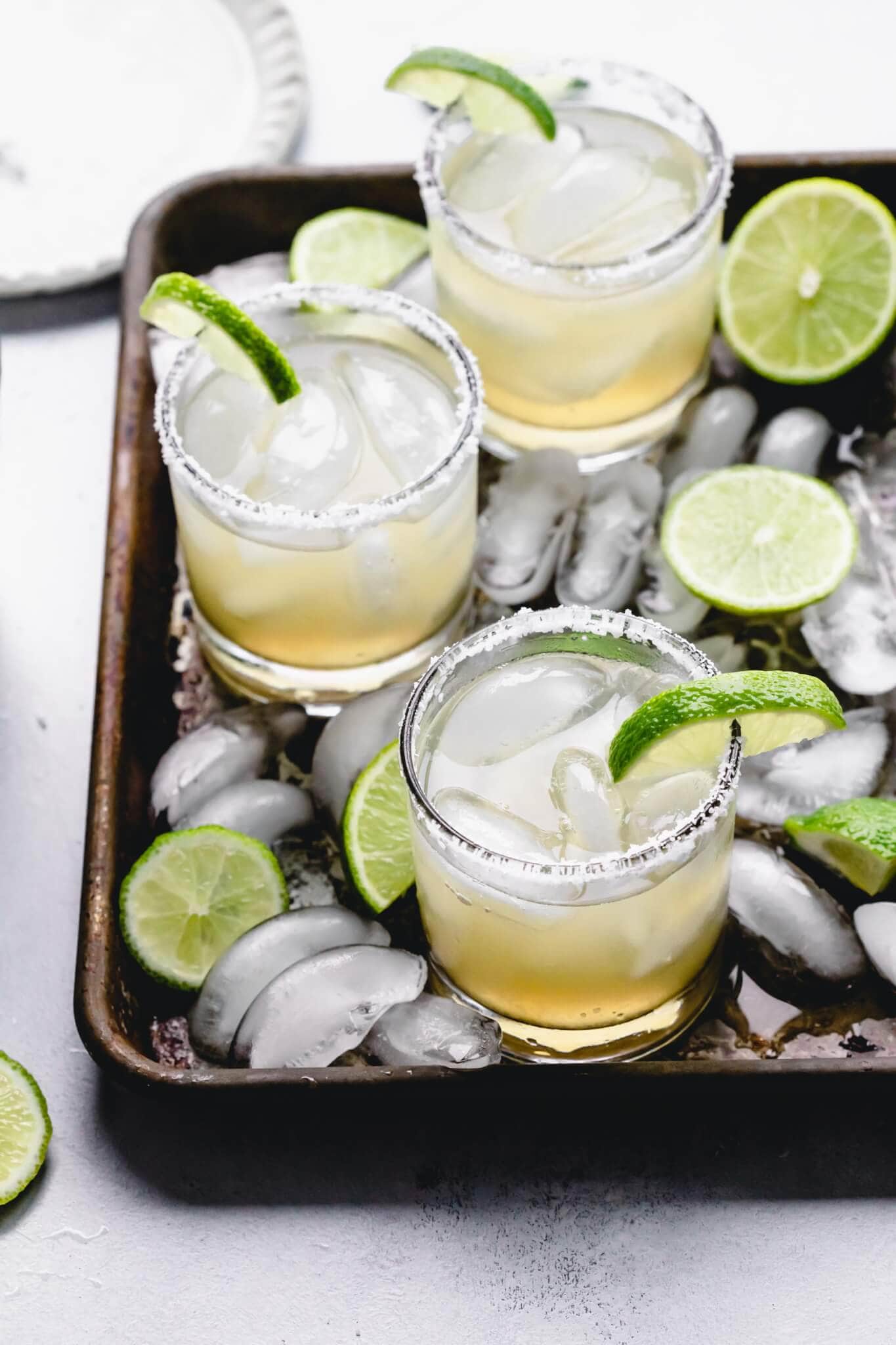 Overhead angle shot of three margaritas on serving tray with ice cubes and lime wedges.