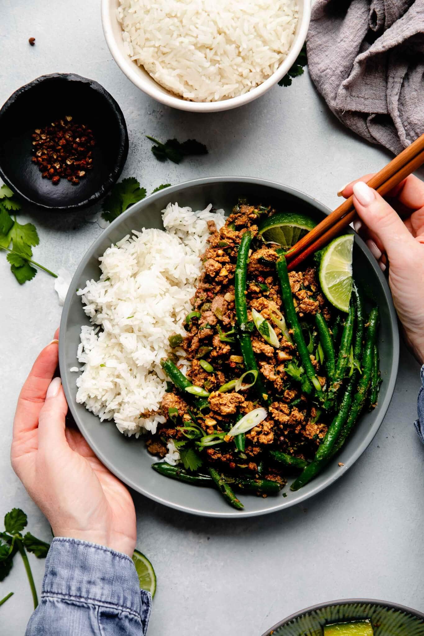 Two hands holding chopsticks and bowl of lamb curry next to bowl of rice.