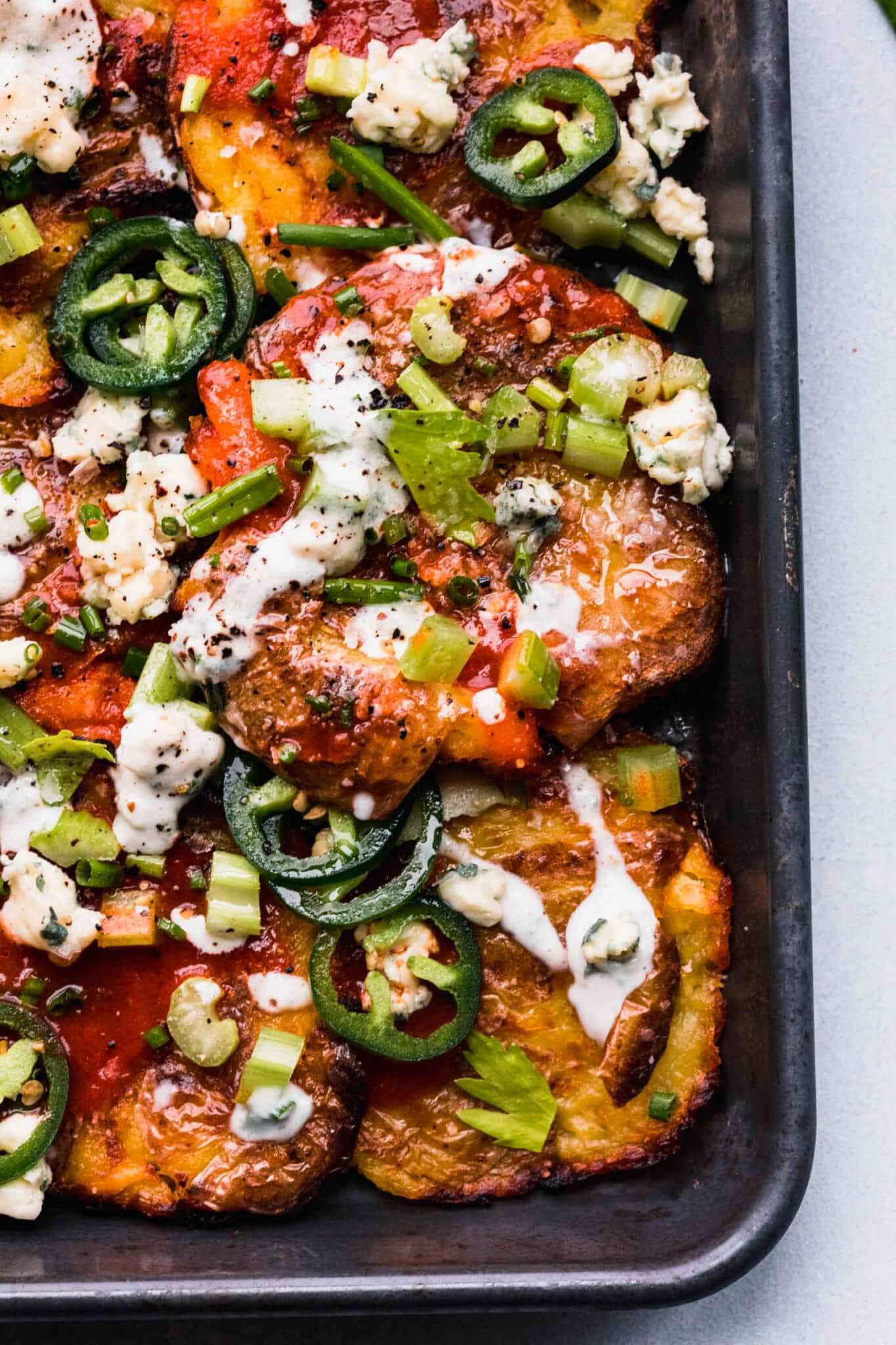 Overhead shot of smashed potatoes on baking sheet topped with blue cheese, hot sauce, jalapenos and chives.