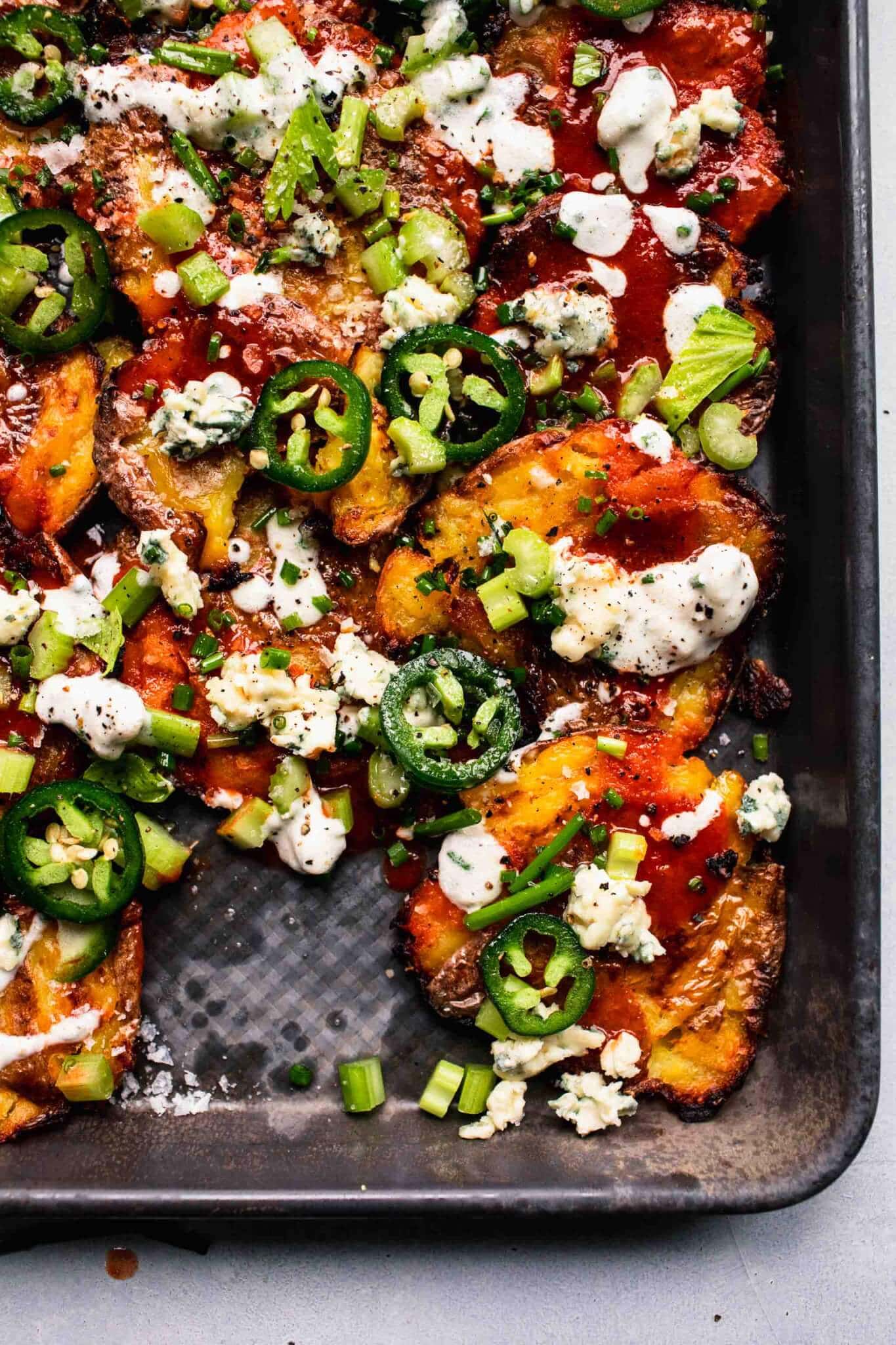 Overhead shot of smashed potatoes on baking sheet topped with blue cheese, hot sauce, jalapenos and chives.