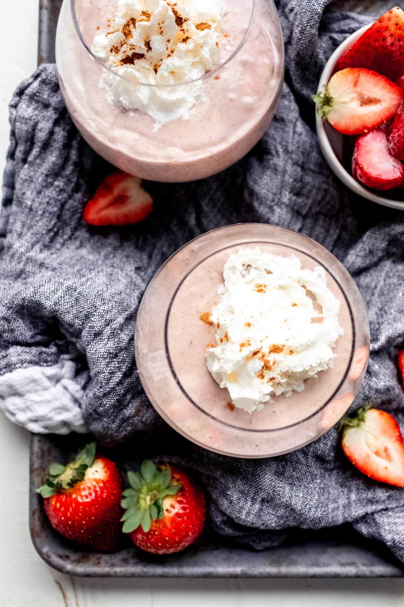 Overhead shot of two smoothies on tray with grey napkin and small bowl of strawberries.