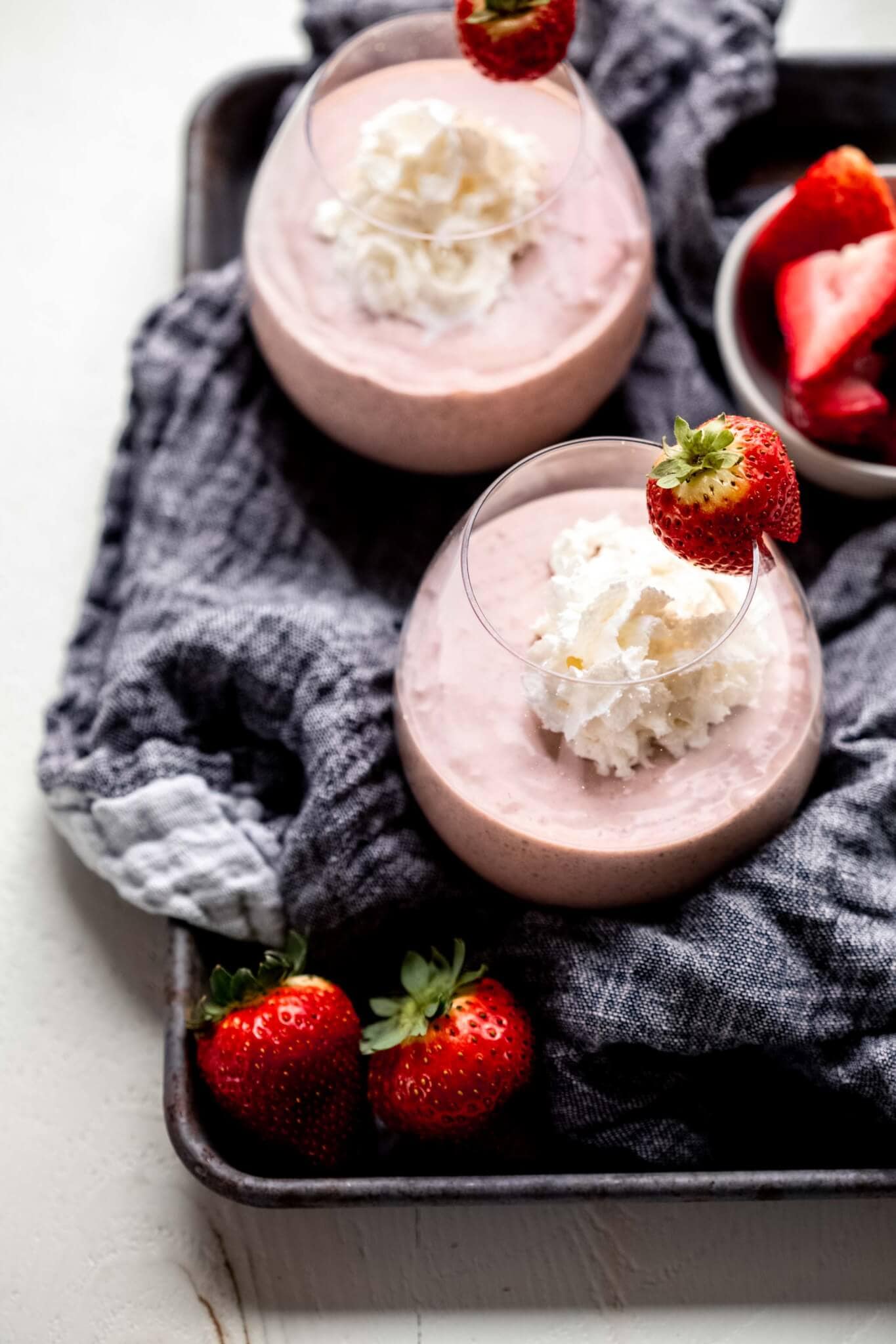 Side view of two healthy strawberry smoothies on tray with grey towel and strawberries scattered about.