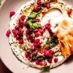 Overhead shot of tahini yogurt sauce in bowl topped with pomegranate seeds and with pita dipped into it.