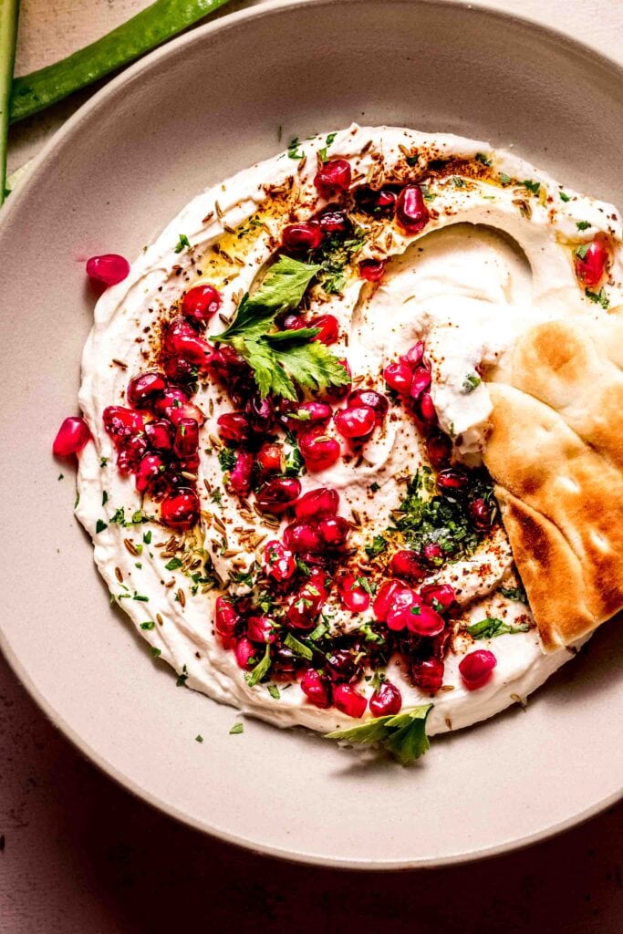 Overhead shot of tahini yogurt sauce in bowl topped with pomegranate seeds and with pita dipped into it.