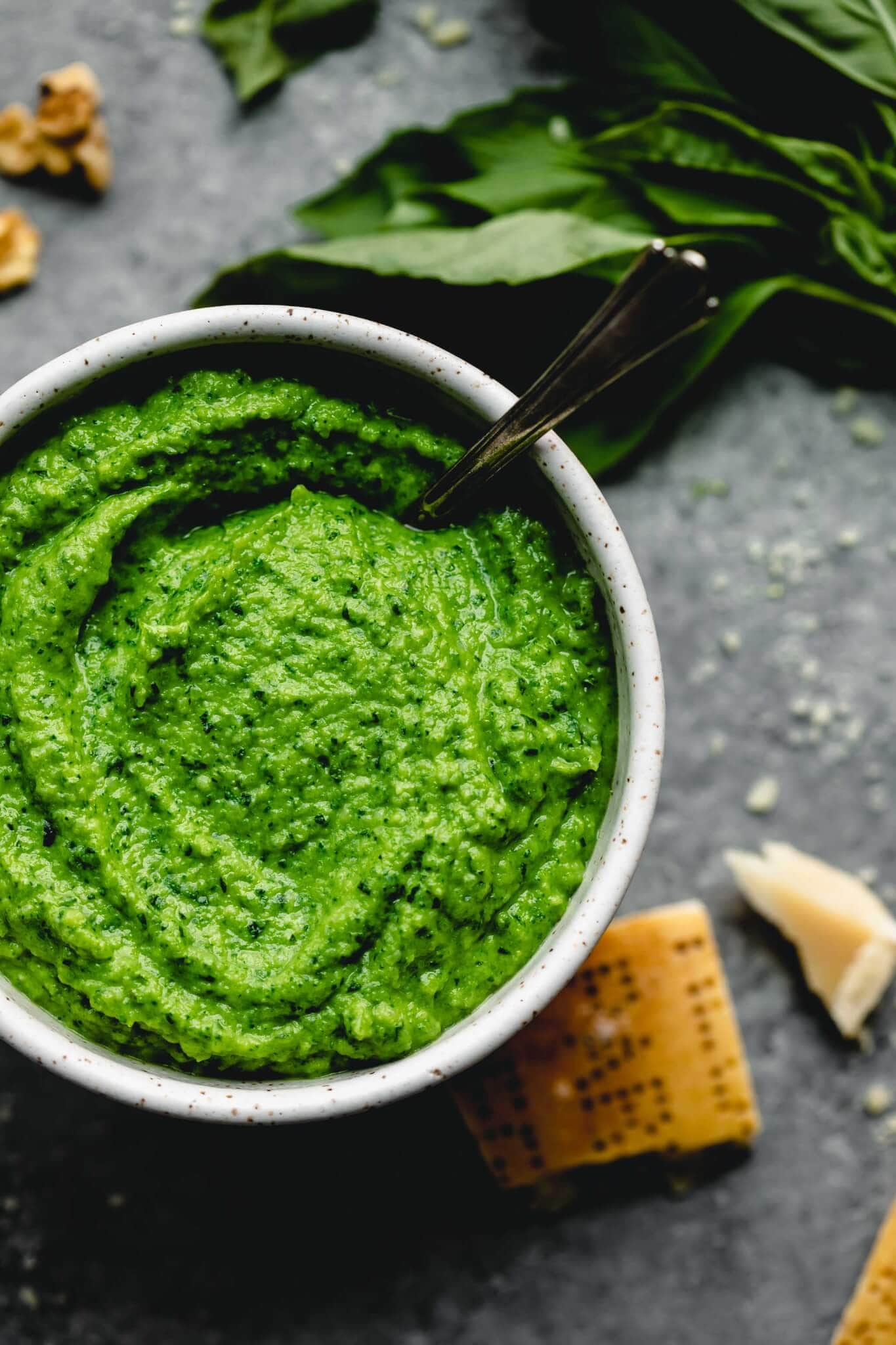 Broccoli pesto in bowl with spoon next to basil leaves and wedge of parmesan.