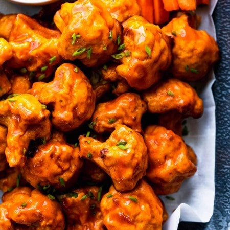 Overhead shot of baked buffalo cauliflower bites in serving dish next to small bowl of blue cheese and carrots & celery sticks.