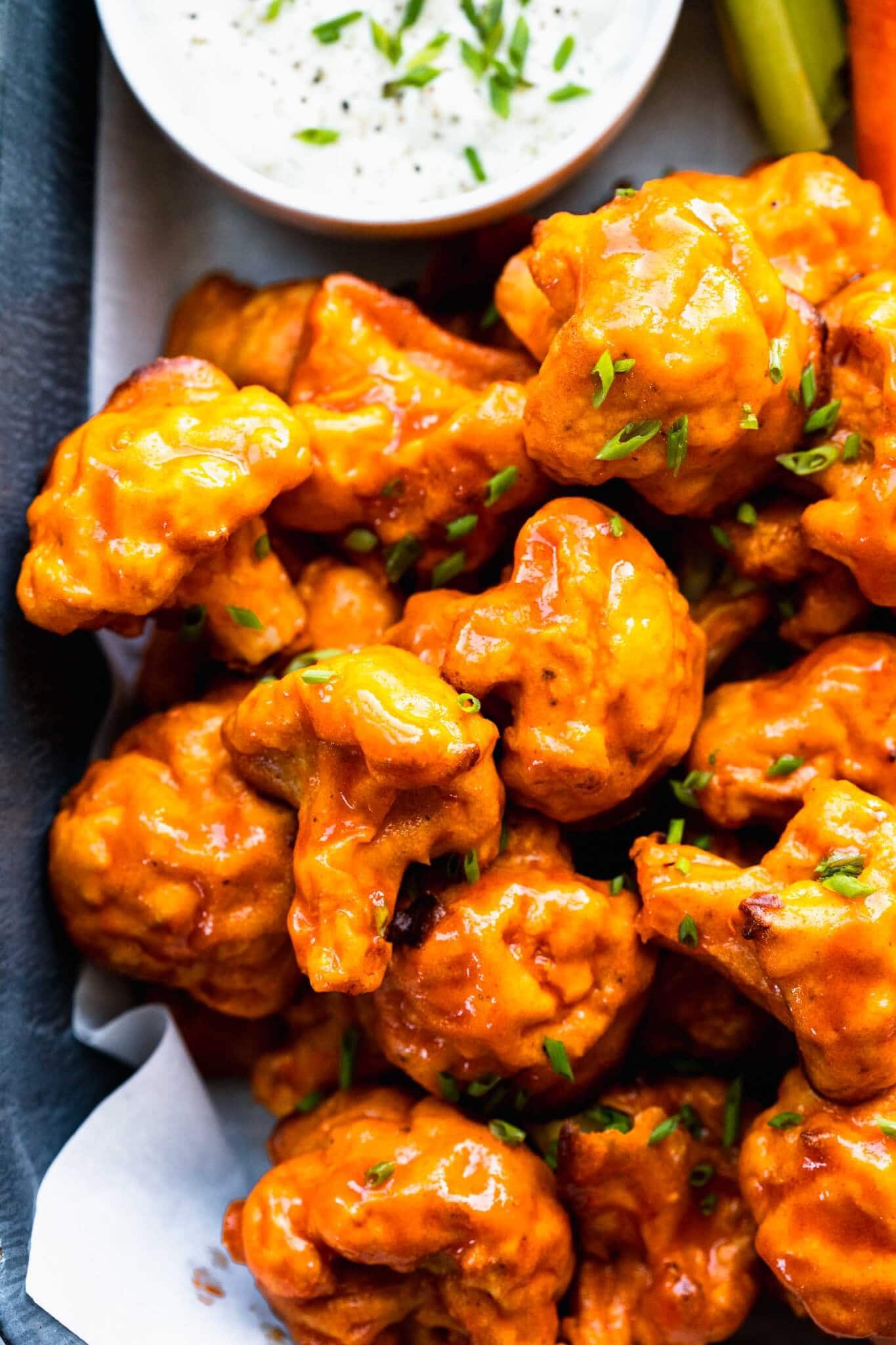 Overhead shot of baked buffalo cauliflower bites in serving dish next to small bowl of blue cheese and carrots & celery sticks.