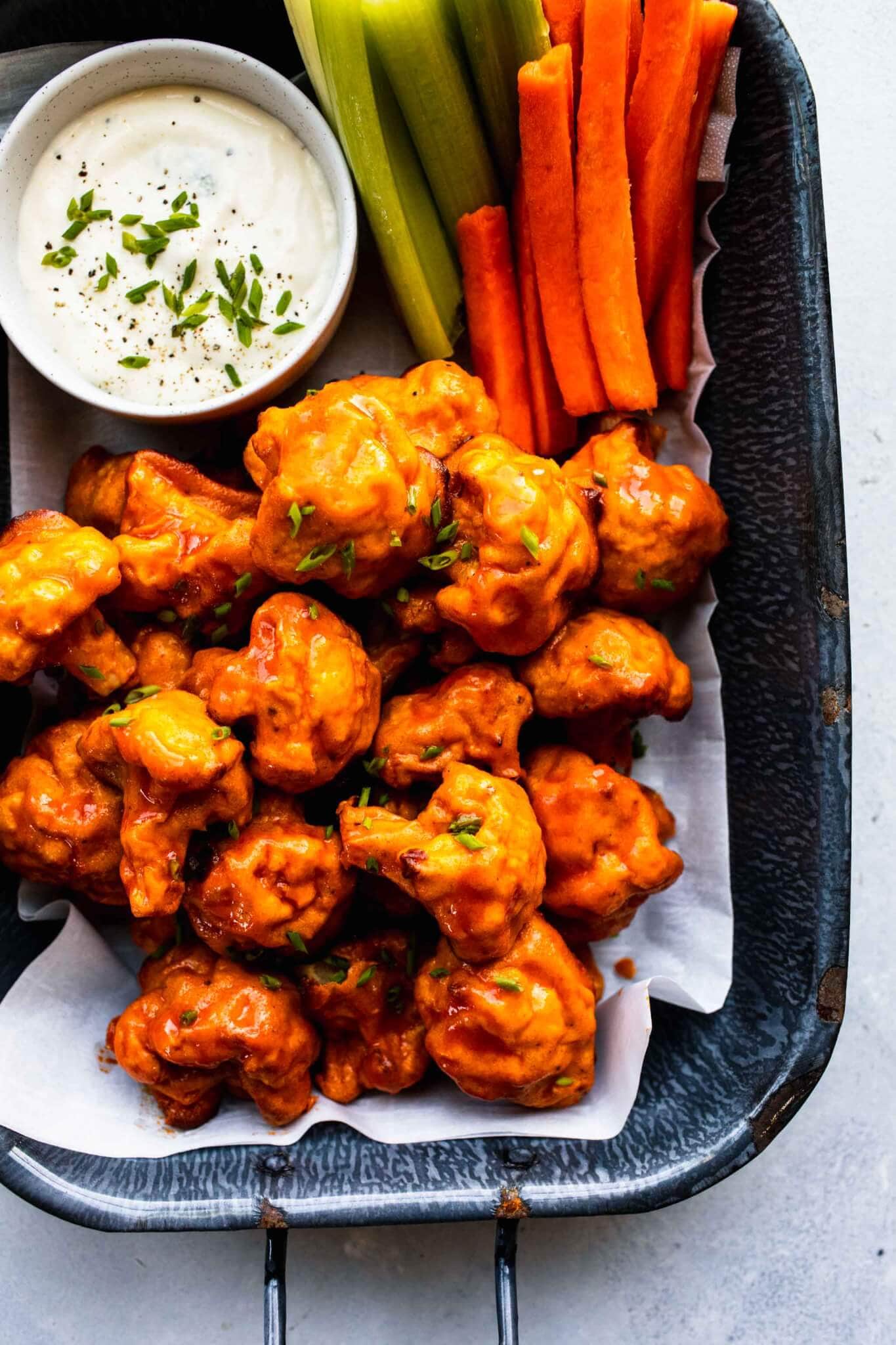 Overhead shot of baked buffalo cauliflower bites in serving dish next to small bowl of blue cheese and carrots & celery sticks.