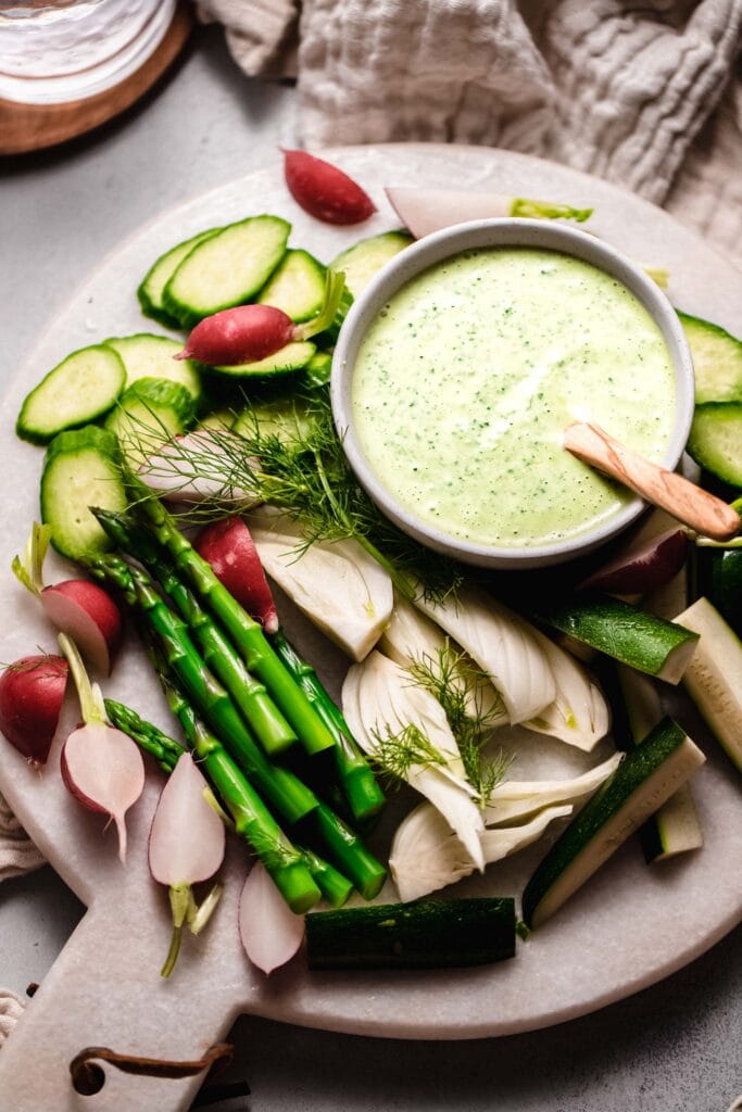 Green goddess dip in bowl with wooden spoon surrounded by veggies.