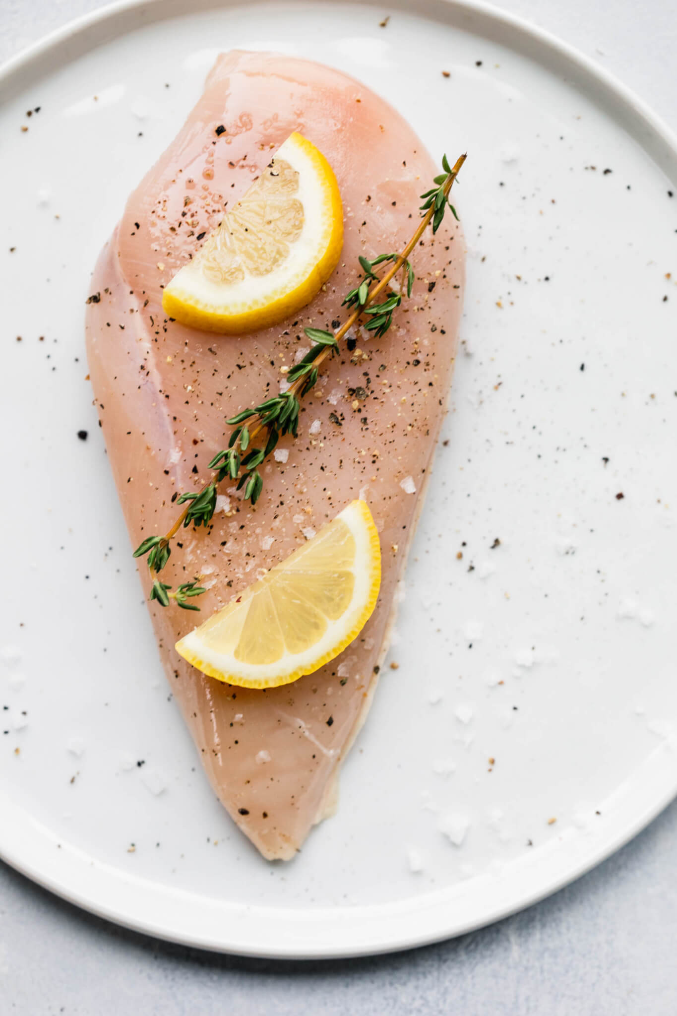 Uncooked chicken breast on small white plated topped with thyme sprigs and lemon slices.