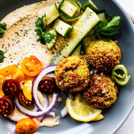 Three air fryer falafel balls in grey bowl with hummus, cucumber and cherry tomatoes.