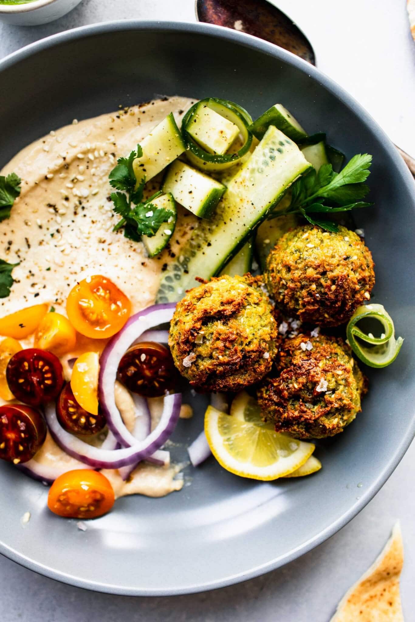 Three air fryer falafel balls in grey bowl with hummus, cucumber and cherry tomatoes.