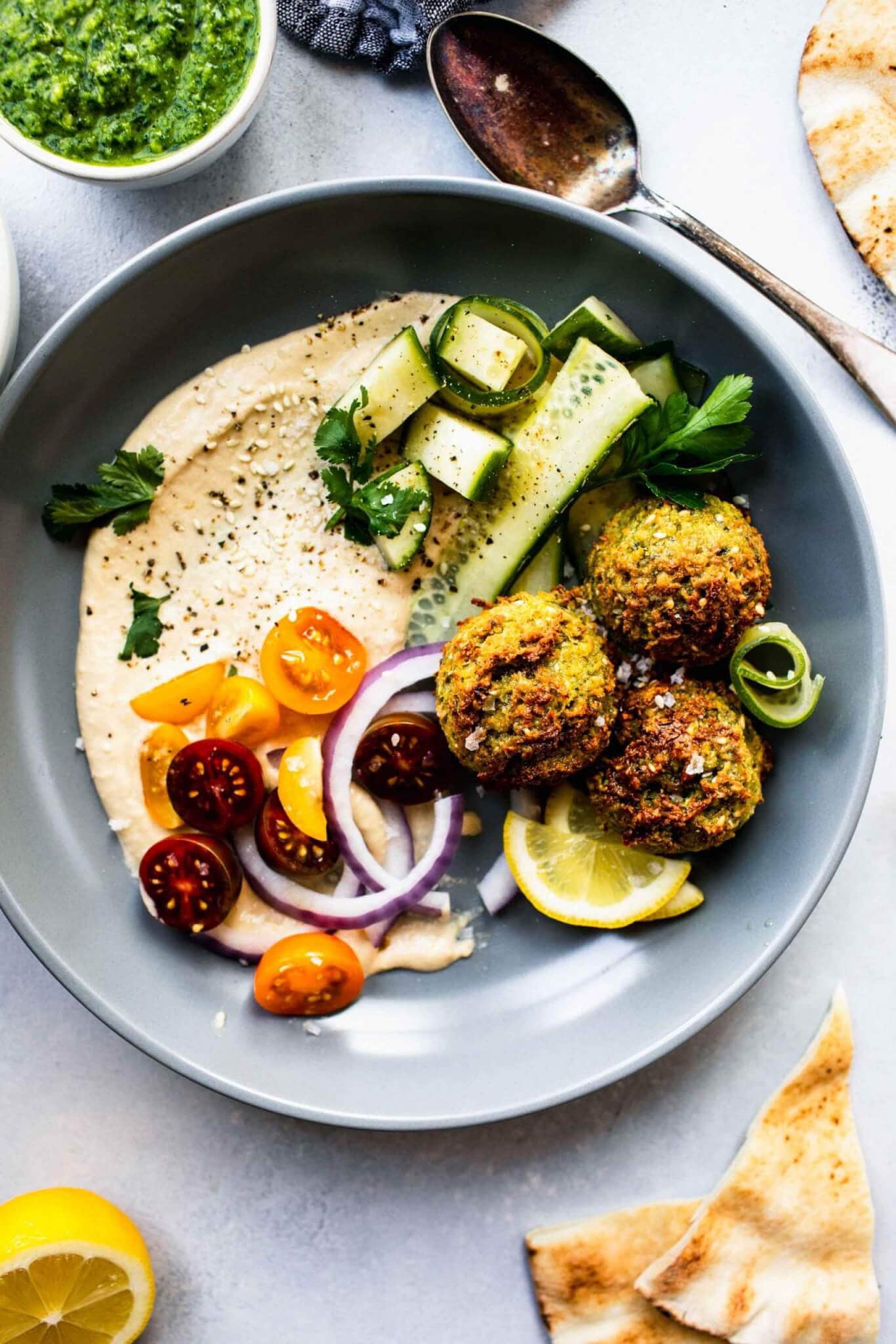 Three air fryer falafel balls in grey bowl with hummus, cucumber and cherry tomatoes.