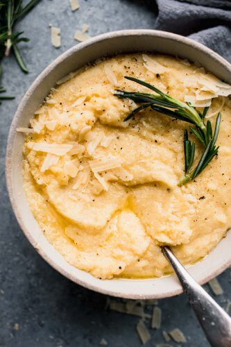 Overhead shot of creamy polenta in white bowl with spoon, topped with parmesan and a sprig of rosemary.