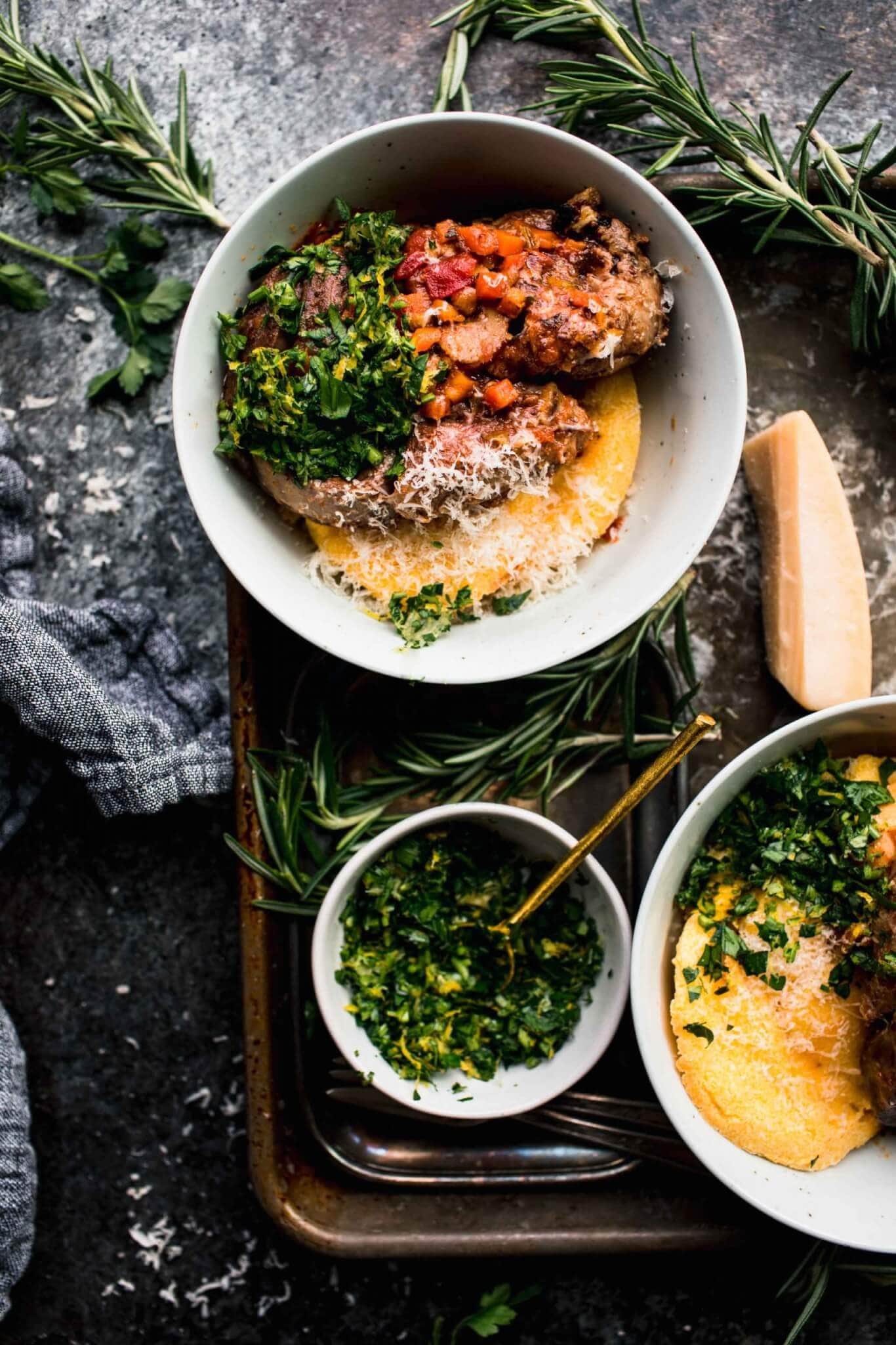 Two bowls of prepared osso buco on top of polenta on serving tray next to sprigs of fresh herbs and bowl of gremolata.