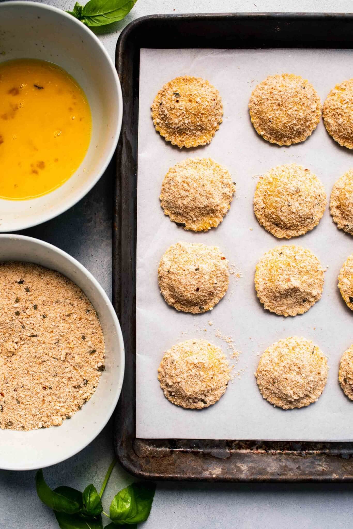 Breaded ravioli on baking sheet before cooking, next to bowl of eggs and bowl of breadcrumbs.