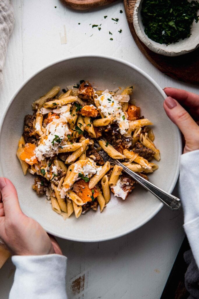 Hands holding bowl of creamy butternut squash pasta. 