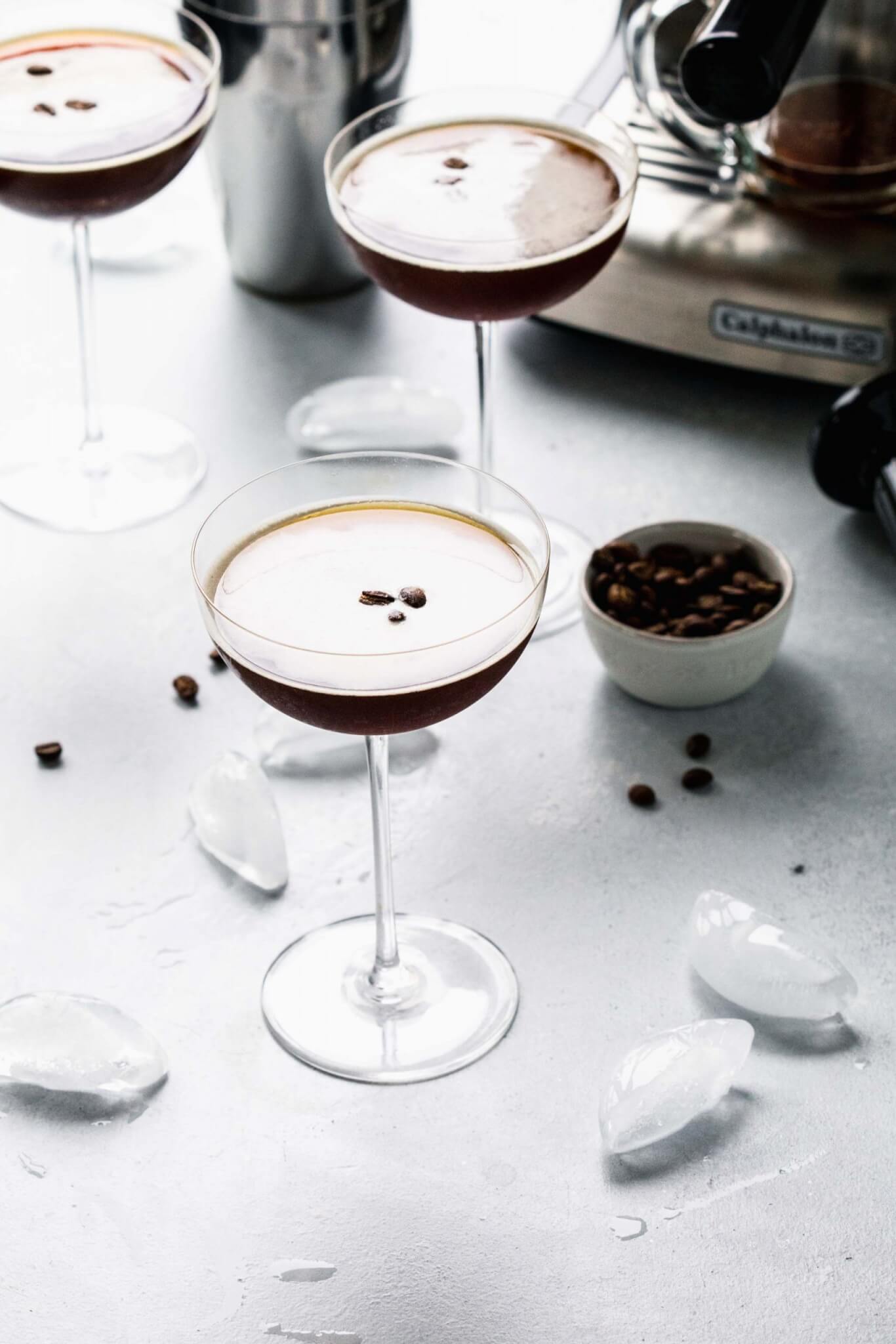Three glasses of espresso martini on counter next to small bowl of coffee beans.