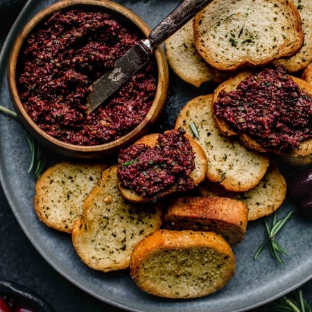 Overhead shot of small bowl of tapenade on grey plate with crostini scattered about.