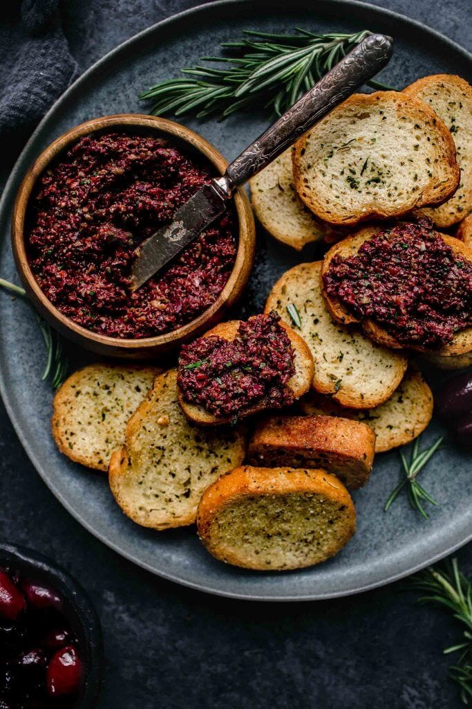 Overhead shot of small bowl of tapenade on grey plate with crostini scattered about.